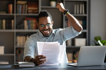 Excited African American man in glasses triumph reading good news in paperwork letter, overjoyed...