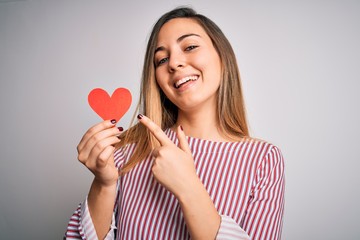 Young beautiful blonde romantic woman holding red paper heart over white background very happy pointing with hand and finger
