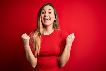 Young beautiful blonde woman with blue eyes wearing casual t-shirt over red background celebrating surprised and amazed for success with arms raised and open eyes. Winner concept.