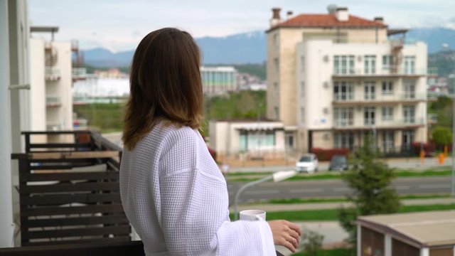 Woman in a bathrobe on a balcony drinks coffee and looks at the street