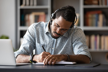 Concentrated African American male student in glasses and wireless headset studying handwriting in...