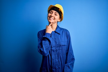 Young beautiful worker woman with blue eyes wearing security helmet and uniform looking confident at the camera with smile with crossed arms and hand raised on chin. Thinking positive.