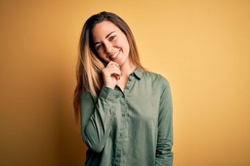 Young beautiful blonde woman with blue eyes wearing green shirt over yellow background looking confident at the camera with smile with crossed arms and hand raised on chin. Thinking positive.