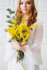 Portrait of a red-haired beautiful girl with a Mimosa in a long white dress