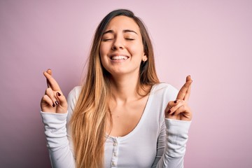 Young beautiful blonde woman with blue eyes wearing white t-shirt over pink background gesturing finger crossed smiling with hope and eyes closed. Luck and superstitious concept.