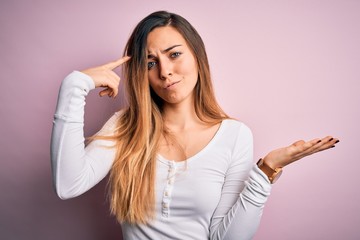 Young beautiful blonde woman with blue eyes wearing white t-shirt over pink background confused and annoyed with open palm showing copy space and pointing finger to forehead. Think about it.
