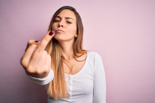 Young beautiful blonde woman with blue eyes wearing white t-shirt over pink background Showing middle finger, impolite and rude fuck off expression