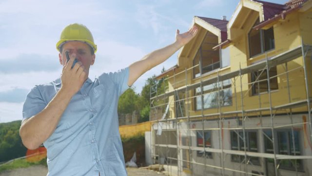 SLOW MOTION, CLOSE UP: Angry Caucasian contractor shouts at his co-worker over the walkie talkie while standing outside of a house under construction. Architect gets into an argument with contractors.