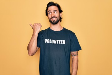 Young handsome hispanic volunteer man wearing volunteering t-shirt as social care smiling with happy face looking and pointing to the side with thumb up.