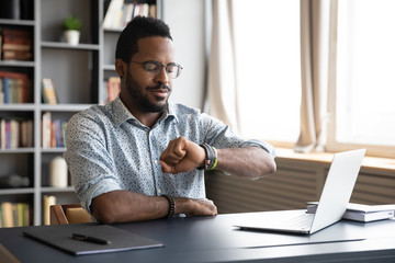 African American young male employee in glasses check time on hand watch waiting for work day to be over, biracial man worker sit at desk use smartwatch at workplace, time management concept - obrazy, fototapety, plakaty