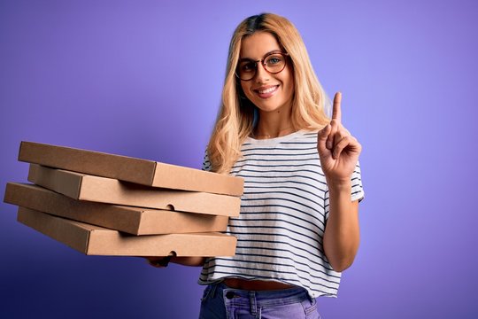 Young beautiful blonde woman holding boxes of italian pizza over isolated purple background surprised with an idea or question pointing finger with happy face, number one