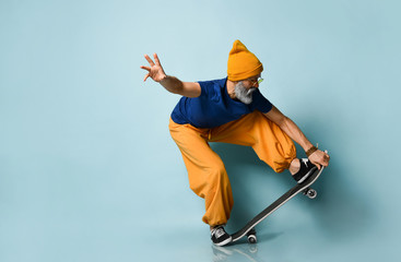 Gray-bearded grandpa in t-shirt, sunglasses, orange pants, hat, gumshoes. Riding black skateboard, posing on blue background