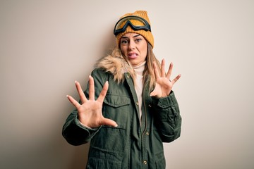 Young brunette skier woman wearing snow clothes and ski goggles over white background afraid and terrified with fear expression stop gesture with hands, shouting in shock. Panic concept.