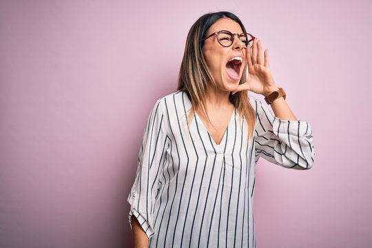Young beautiful woman wearing casual striped t-shirt and glasses over pink background shouting and screaming loud to side with hand on mouth. Communication concept.