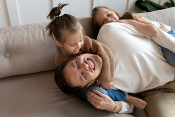 Joyful little sisters cuddling laughing father, resting together on sofa. Playful small daughters...