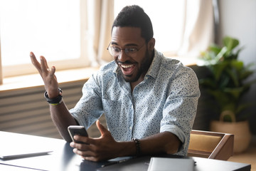 Excited African American man in glasses sit at desk triumph win online lottery on cellphone, overjoyed biracial male in spectacles feel euphoric read good unexpected news or message on smartphone - obrazy, fototapety, plakaty