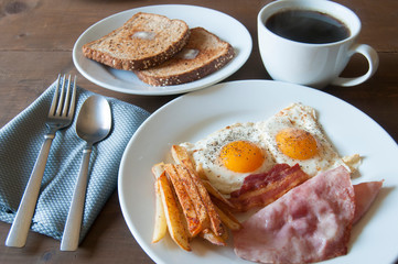 Breakfast on a table with eggs ham bacon fries and toast with butter on the side and black coffee