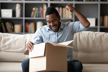 Excited biracial young man sit on couch at home satisfied with Internet order shopping online,...