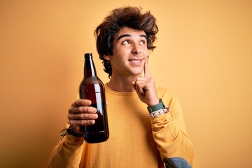 Young handsome man drinking bottle of beer standing over isolated yellow background serious face...