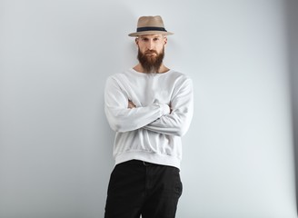 Serious bearded man in hat standing against white wall arms crossed.