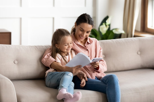 Attractive young mixed race woman nanny cuddling little kid, reading paper book together on comfortable sofa in living room. Happy mother educating small child daughter, enjoying weekend time at home.