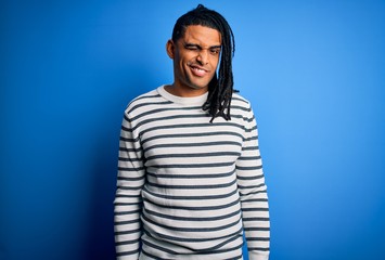 Young handsome african american afro man with dreadlocks wearing casual striped sweater winking...