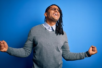Fototapeta premium Young handsome african american man with dreadlocks wearing casual sweater and glasses very happy and excited doing winner gesture with arms raised, smiling and screaming for success. Celebration 