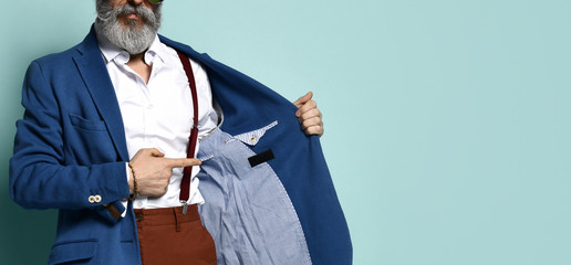 Cropped photo of man in white shirt, sunglasses, brown pants, suspenders, He pointing at label on the inside of his jacket, posing on blue background