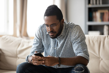 Focused african American man in glasses sit on couch in living room browsing internet on...