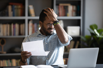 Frustrated biracial male student sit at desk feel distressed with bad news in paper letter, unhappy African American man stressed by dismissal notice or negative reply in post correspondence or notice - obrazy, fototapety, plakaty