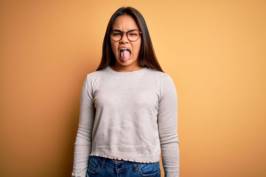 Young beautiful asian girl wearing casual sweater and glasses over yellow background sticking tongue out happy with funny expression. Emotion concept.