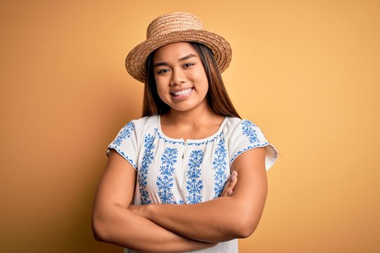 Young beautiful asian girl wearing casual t-shirt and hat standing over yellow background happy face smiling with crossed arms looking at the camera. Positive person.