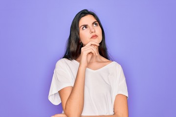 Young beautiful brunette woman wearing casual white t-shirt over purple background with hand on chin thinking about question, pensive expression. Smiling with thoughtful face. Doubt concept.