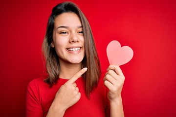 Young beautiful brunette romantic girl holding red paper heart shape over isolated background very happy pointing with hand and finger