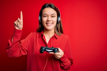 Young beautiful brunette gamer girl playing video game using joystick and headphones surprised with an idea or question pointing finger with happy face, number one © Krakenimages.com