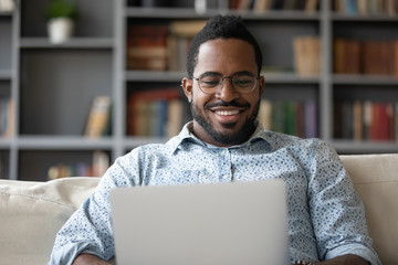 Smiling biracial millennial man in glasses sit relax on couch in living room browse wireless...
