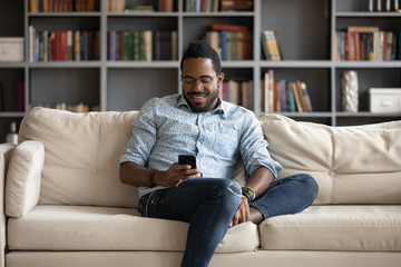 Smiling african American man sit relax on comfortable couch at home using modern cellphone, happy biracial millennial male rest on sofa in living room texting messaging on smartphone gadget - obrazy, fototapety, plakaty