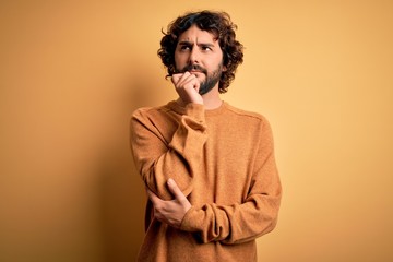 Young handsome man with beard wearing casual sweater standing over yellow background with hand on chin thinking about question, pensive expression. Smiling and thoughtful face. Doubt concept.