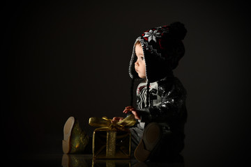 Fototapeta na wymiar Little child in gray hat, suit and boots. She looking up, holding golden gift box, sitting on floor. Black background. Close up