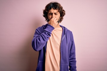 Young handsome sporty man with beard wearing casual sweatshirt over pink background smelling something stinky and disgusting, intolerable smell, holding breath with fingers on nose. Bad smell