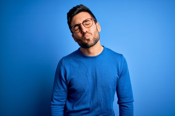 Young handsome man with beard wearing casual sweater and glasses over blue background looking at the camera blowing a kiss on air being lovely and sexy. Love expression.