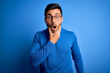 Young handsome man with beard wearing casual sweater and glasses over blue background Looking fascinated with disbelief, surprise and amazed expression with hands on chin