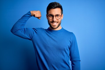 Young handsome man with beard wearing casual sweater and glasses over blue background Strong person...