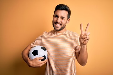 Handsome player man with beard playing soccer holding footballl ball over yellow background smiling with happy face winking at the camera doing victory sign with fingers. Number two.