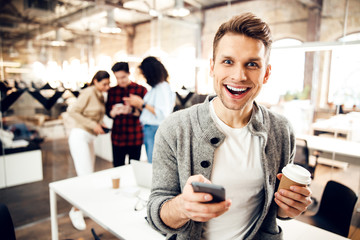 Cheerful young man holding smartphone and cup of coffee