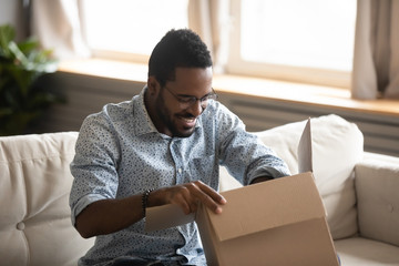 Excited biracial young man sit on couch at home unpack cardboard box with Internet order, happy...