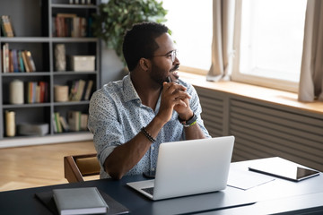 Smiling African American man in glasses sit at desk distracted from computer work look in distance...