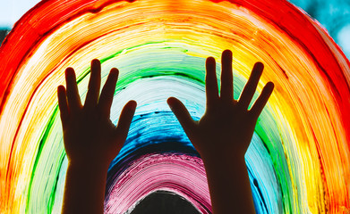 Child hands touch painting rainbow on window.