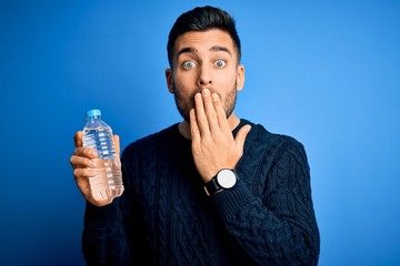 Young handsome man drinking bottle of water to refeshment over blue background cover mouth with hand shocked with shame for mistake, expression of fear, scared in silence, secret concept