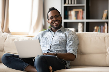 Portrait of smiling biracial young man in glasses sit relax on couch at home using laptop gadget,...